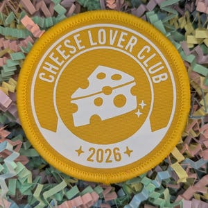 May include: A circular, yellow patch with the words "CHEESE LOVER CLUB" in white, surrounding a white cheese graphic. The patch also features the year "2026" and two small star graphics. The patch is edged with a yellow border.