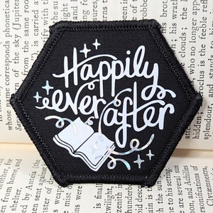 May include: A black hexagonal patch with white text that reads "Happily ever after". The text is styled with a whimsical font. A white open book with a glittery effect is in the centre of the patch.