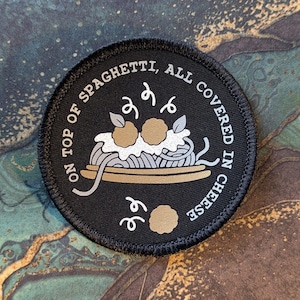May include: A circular black embroidered patch featuring a graphic of spaghetti with meatballs and cheese. The text reads: ON TOP OF SPAGHETTI, ALL COVERED IN CHEESE. The design includes white and tan colours, with a stitched border edge and a flat surface.