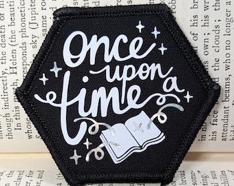 Peut inclure: Un écusson hexagonal noir avec des coutures blanches autour des bords. L'écusson présente un motif blanc d'un livre ouvert avec des étoiles et le texte "Once upon a time" dans une police de caractères fantaisiste.