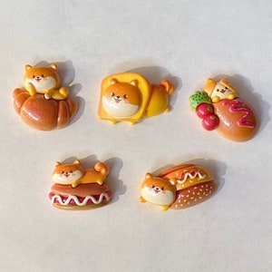 May include: Five colorful clay charms featuring cartoon dogs in various poses. The charms are shaped like a croissant, a hot dog, a loaf of bread, and a lion.