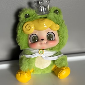 May include: A plush doll dressed in a green frog costume with a clear plastic crown. The doll has a yellow wig, rosy cheeks, and large green eyes. The frog costume has yellow feet and a white cape.