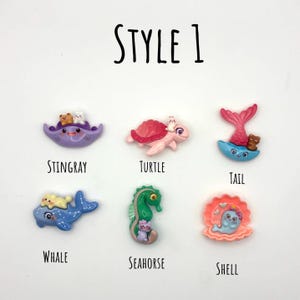 Puede incluir: Una colección de seis figuras en miniatura coloridas con temática oceánica. Las figuras incluyen una manta raya morada, una tortuga rosa, una cola de sirena, una ballena azul, un caballito de mar verde y una concha. El texto "STYLE 1" está en la parte superior.