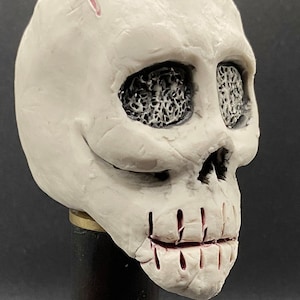 May include: A white ceramic skull with detailed eye sockets and a cracked lip. The skull is mounted on a black wooden base. "Skull 1" is written below the base.