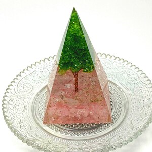 May include: A clear pyramid with a green tree inside, set on a clear glass plate with a decorative pattern. The tree is made of green crystals and has a copper wire trunk. The base of the pyramid is filled with pink crystals.