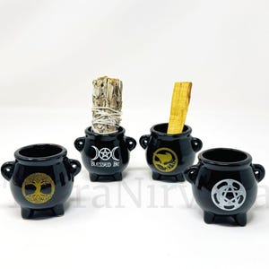 May include: Four small black ceramic cauldron incense holders with gold and silver symbols. One cauldron holds a bundle of sage, another a piece of palo santo. The text "Blessed Be" is on one cauldron.