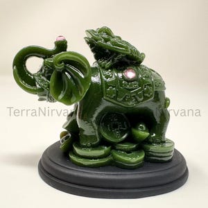 May include: A jade green elephant figurine with a dragon on its back, set on a black base. The elephant has its trunk raised and is adorned with pink accents. The figurine is approximately 10 cm tall.