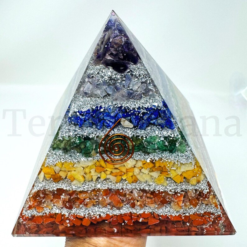May include: A layered pyramid sculpture with a clear exterior. The pyramid is filled with colorful gemstones and silver flakes, arranged in horizontal bands of red, orange, yellow, green, blue, and purple. A copper spiral sits on the front.