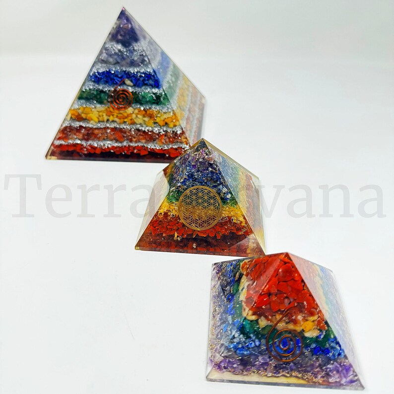May include: Three colorful pyramid-shaped orgone energy generators. Each pyramid features layers of vibrant gemstones in a rainbow spectrum, with a metallic symbol on the front. The text "Terra vana" is visible in the background.