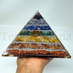 May include: A clear pyramid-shaped object with layers of colorful stones and a copper spiral in the center. The layers include red, orange, yellow, green, blue, and purple stones, separated by silver glitter. The pyramid is held by a hand.