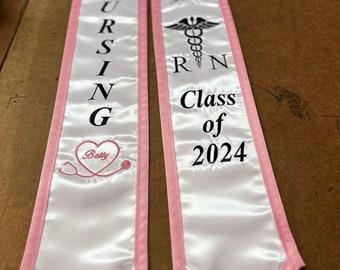 May include: Two white satin graduation sashes with pink trim. One sash says "NURSING" with a stethoscope and a heart that says "Betty". The other sash says "Salina Gomez RN Class of 2024" with a caduceus symbol.