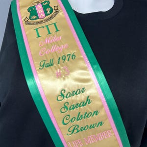 May include: A gold satin sash with green and pink trim. The sash features embroidered text and a crest. Text includes "ΓΠ", "Miles College Fall 1976", and names. The sash is draped over a black garment.