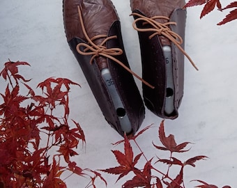 May include: A pair of brown leather shoes with laces. The shoes have a brown leather upper and a brown leather sole. The shoes are on a white surface with red leaves.