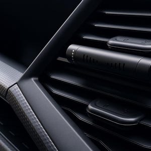 May include: A close-up of a car's air vent featuring a black cylindrical air freshener. The vent is black with horizontal slats and two rectangular control buttons. The interior trim has a carbon fibre pattern.