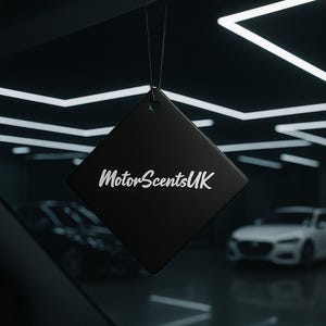 May include: A black, diamond-shaped car air freshener with the text "MotorScentsUK" in white. The freshener is suspended by a black string. The background shows a car interior and a showroom.