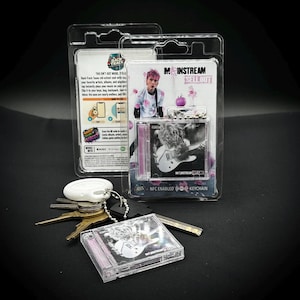 Peut inclure: Un emballage en plastique transparent contenant un porte-clés avec un mini boîtier de CD représentant un musicien jouant de la guitare. L'emballage porte l'inscription "Mainstream Sellout" et "NFC Enabled Keychain". Un jeu de clés et un porte-clés blanc sont au premier plan.