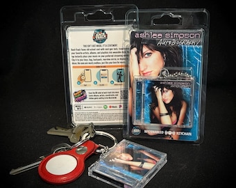 May include: An Ashlee Simpson Autobiography NFC-enabled keychain in clear plastic packaging, a loose matching keychain, and a red leather-like key fob with a silver metal ring and attached keys. The packaging features the album cover art and text.