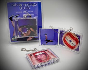 May include: Clear plastic keychain featuring album art from Olivia Rodrigo's "GUTS." The keychain has a square shape with a silver chain and a small image inside. The packaging includes the text "olivia rodrigo GUTS" and "NFC ENABLED KEYCHAIN."