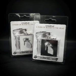 May include: Two keychains featuring the album art from Coldplay's "A Rush of Blood to the Head." Each keychain has a small, square image of the album cover in a clear plastic case. The packaging includes the band's name and album title.