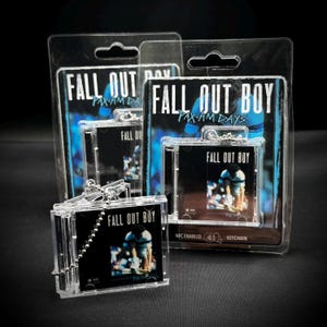 May include: Clear plastic keychain featuring a miniature CD case with the text "FALL OUT BOY" and "PAX-AM DAYS". The CD case has a photo of a person on the cover. The keychain is packaged in a clear plastic blister pack.