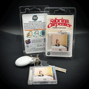 Może przedstawiać: Przezroczysty plastikowy brelok do kluczy z miniaturowym etui na CD ze zdjęciem Sabriny Carpenter. Brelok jest przymocowany do zestawu kluczy. Na opakowaniu widnieje napis "Sabrina Carpenter emails i can't send".
