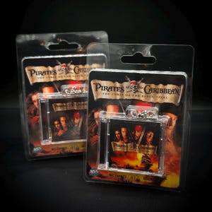 Puede incluir: Dos llaveros de Piratas del Caribe en embalaje de plástico transparente. Cada llavero presenta un estuche de CD en miniatura con ilustraciones de la película y el título. El embalaje incluye el título de la película e imágenes de personajes.