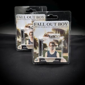 Puede incluir: Dos llaveros de plástico transparente con la portada del álbum "American Beauty/American Psycho" de Fall Out Boy. Cada llavero muestra una foto de una persona con pintura facial. El embalaje incluye el nombre de la banda y el título del álbum.