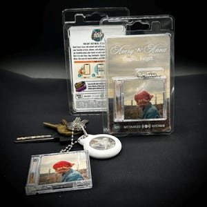 Peut inclure: Un porte-clés en plastique transparent avec un petit design de boîtier de CD, présentant une image d'une personne portant un couvre-chef rouge. Le porte-clés est attaché à une lumière blanche de forme ovale. L'emballage indique "Avery & Anna" et "forgive, forget."
