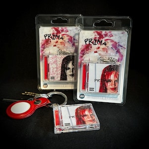 May include: Two clear plastic blister packs containing miniature square keychains with the text PRIMA and an image of a person with red hair. A third loose keychain sits in front, alongside a red circular tag attached to a metal key ring.
