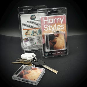 May include: A clear plastic keychain featuring a miniature CD case with a photo of Harry Styles. The packaging reads "Harry Styles" and "NFC ENABLED KEYCHAIN". A set of keys and a white key fob are also visible.