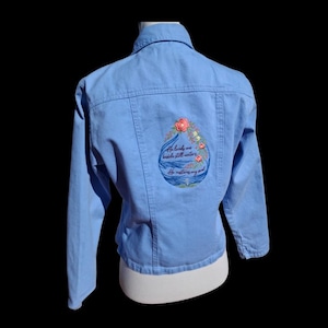 May include: A light blue denim jacket featuring an embroidered design on the back. The embroidery depicts a water droplet shape with flowers, waves, and the text: He leads me beside still waters, He restores my soul. The jacket has a collar.