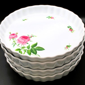 May include: A stack of four round white ceramic quiche dishes with fluted edges. The top dish features a floral pattern with pink roses, green leaves, and small rosebuds scattered across the white surface against a plain black background.