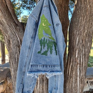 May include: Light blue denim jacket with a green wolf and forest design on the back. The jacket has a frayed hem on the sleeves and button closures. The wolf design includes a crescent moon and stars.