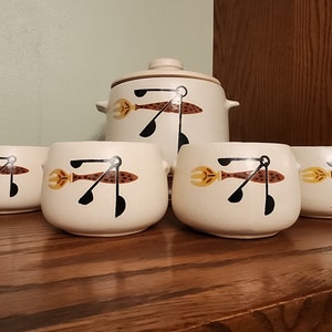 May include: A cream-colored ceramic soup tureen with a lid and four matching bowls. Each piece features a repeating design of a brown fish, yellow fork, and black spoon. The set is displayed on a wooden surface.