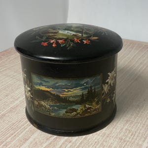 May include: A round, black wooden box with a lid. The lid features a painted landscape with red flowers. The sides of the box display a scenic landscape with a lake, trees, and mountains. White flowers are painted on the sides.