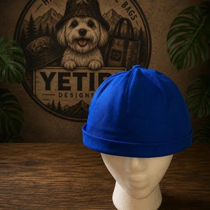 Peut inclure: Une toque de marin bleu roi uni avec une calotte arrondie et un bord épais replié. Le chapeau est présenté sur une tête de mannequin blanche, mettant en valeur son design simple sans bordure et sa construction en tissu lisse sans motifs.