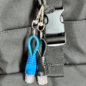Pode incluir: Close-up de uma mochila preta com zíper e fivela. Dois chaveiros de cabo RJ45 pendurados no zíper. Um é azul, o outro é preto. Ambos têm conectores transparentes com fios visíveis.