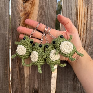 May include: Four green crochet frog keychains with white bellies and black eyes are displayed. The keychains are attached to silver key rings. The frogs are held in front of a wooden fence.
