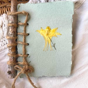 Puede incluir: Un diario hecho a mano con una cubierta verde claro y una flor amarilla prensada. El lomo es una pequeña rama de madera envuelta con hilo. El diario está sobre una superficie blanca.