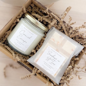 May include: A gift box filled with brown crinkle paper contains a white candle with a gold lid and a cream-colored bar of soap. The candle label reads "Cornish Escape" with the scent "Stone Driftwood & Musk". The soap label reads "Sundays in St Ives" with the scent "Creamy Sandalwood & Vanilla Bean".