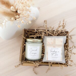 May include: A gift box filled with brown paper shreds contains a white candle with a gold lid and a white bar of soap. The candle label reads "Cornish Escape" and the soap label reads "Sundays in Salt Trees".