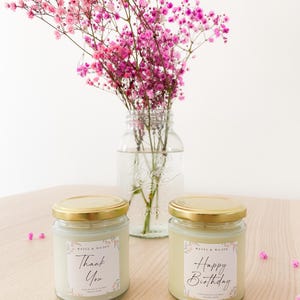 May include: Two glass candles with gold lids and white labels featuring pink floral accents. One label reads "Thank You" and the other "Happy Birthday" in black script. Both include the text "WAVES & WILDER" and "SOY WAX CANDLE".