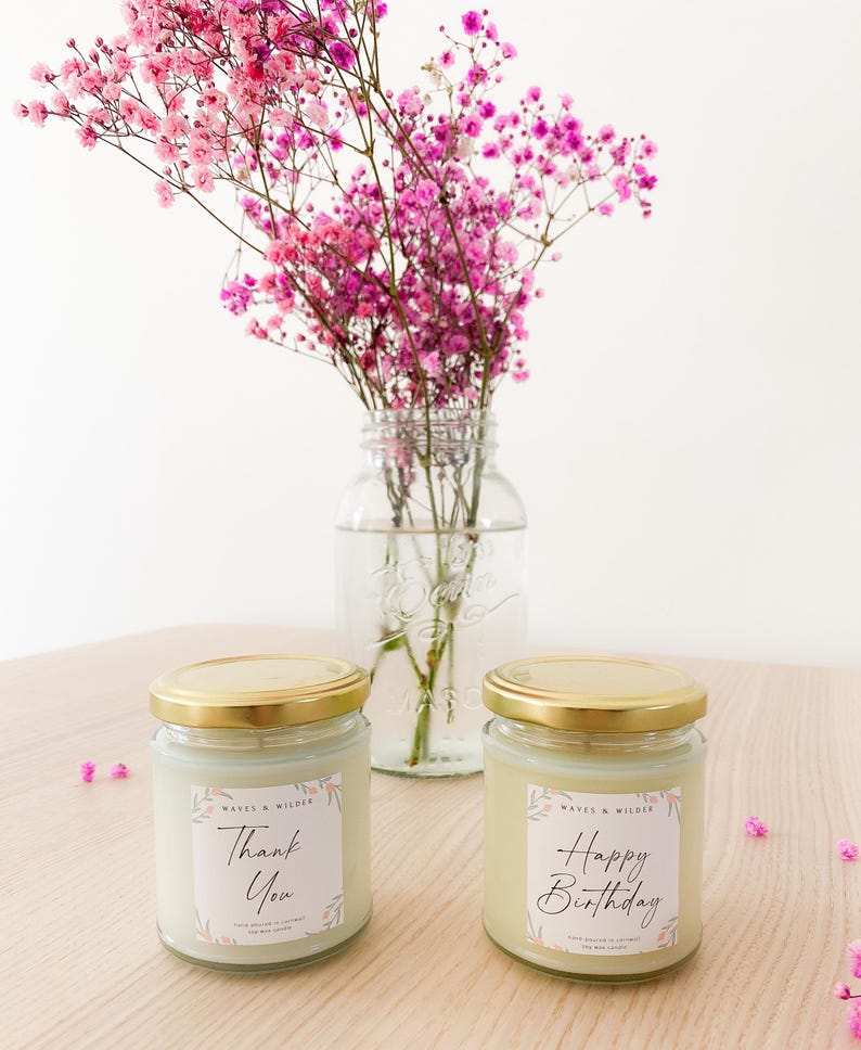 May include: Two glass candles with gold lids and white labels featuring pink floral accents. One label reads "Thank You" and the other "Happy Birthday" in black script. Both include the text "WAVES & WILDER" and "SOY WAX CANDLE".