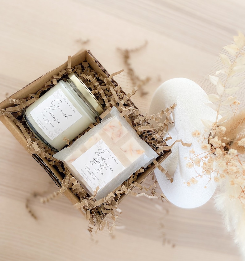 May include: A gift box filled with brown crinkle paper contains a white candle with the text "Cornish Escape" and a white soap bar with the text "Sundays in Spitios".
