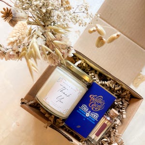 May include: A cardboard gift box containing a glass candle with a Thank You label, a blue Willie's Cacao Milk of the Gods chocolate bar, and a small glass vial of matches with a cork stopper, all nestled in paper filler with dried flowers.