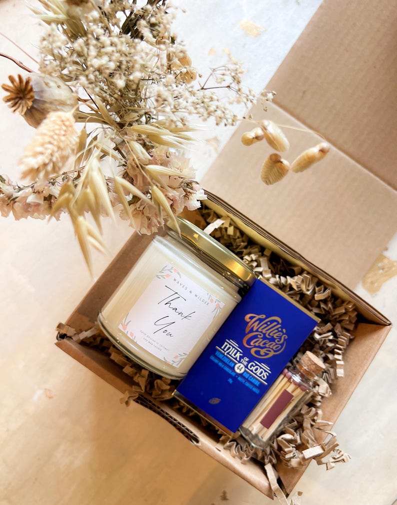 May include: A cardboard gift box containing a glass candle with a Thank You label, a blue Willie's Cacao Milk of the Gods chocolate bar, and a small glass vial of matches with a cork stopper, all nestled in paper filler with dried flowers.