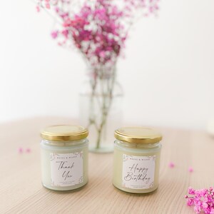 May include: Two glass jars with gold lids containing light-colored wax candles. Each jar features a white square label with floral accents and black text. One label reads "Thank You" and the other reads "Happy Birthday" with "WAVES & WILDER" above.
