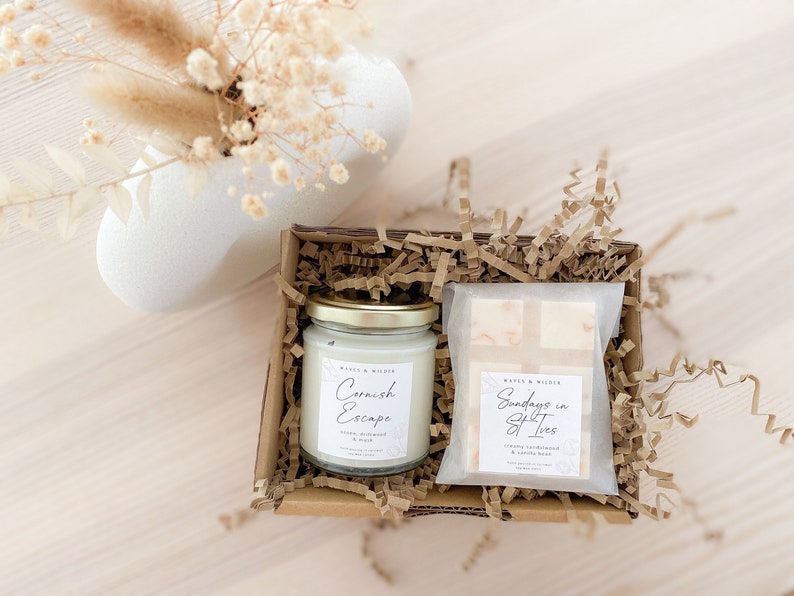 May include: A gift box filled with brown paper shreds contains a white candle with a gold lid and a white bar of soap. The candle label reads "Cornish Escape" and the soap label reads "Sundays in Salt Trees".