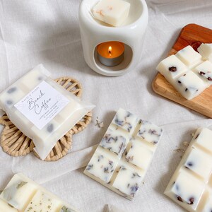May include: A set of wax melts in various scents, including Beach Coffee, Lavender, and a floral scent. The wax melts are packaged in white paper with a black label. The wax melts are displayed on a white surface with a white ceramic wax warmer.