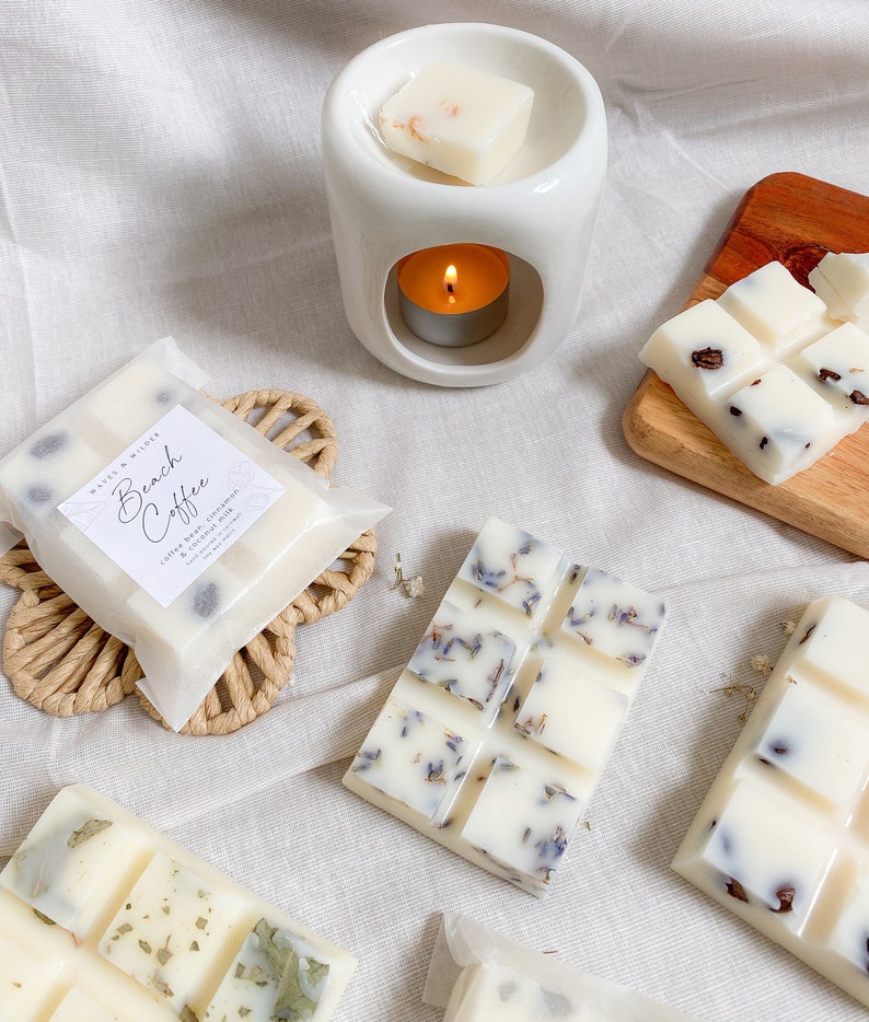 May include: A set of wax melts in various scents, including Beach Coffee, Lavender, and a floral scent. The wax melts are packaged in white paper with a black label. The wax melts are displayed on a white surface with a white ceramic wax warmer.